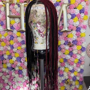 Könnte beinhalten: Eine lange geflochtene Perücke mit schwarzen und burgunderfarbenen Zöpfen. Die Perücke wird auf einem Schaufensterpuppenkopf auf einem Ständer präsentiert. Der Hintergrund ist eine Wand aus bunten Kunstrosen.