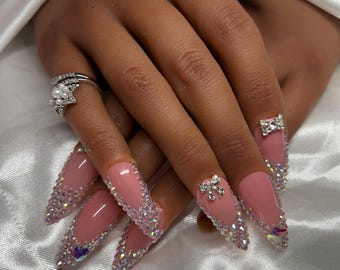 May include: Close-up of hands with long, pink, stiletto-shaped nails decorated with rhinestones and small bows. The nails are adorned with a sparkling, iridescent design.