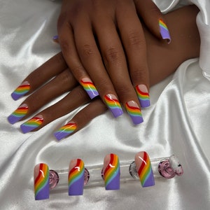 Pode incluir: Unhas artificiais quadradas com um design de arco-íris. As unhas apresentam uma base lavanda com um padrão de arco-íris curvo em vermelho, laranja, amarelo, verde e azul. Um conjunto de unhas de substituição também é exibido.