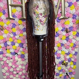 Könnte beinhalten: Eine lange, dunkelbraune Perücke mit Kunst-Locs wird auf einem Schaufensterkopf präsentiert. Die Perücke befindet sich vor einem Hintergrund aus bunten Kunstrosen in Rosa, Gelb und Weiß. Goldene Schmetterlinge und die Buchstaben "HM" sind ebenfalls zu sehen.