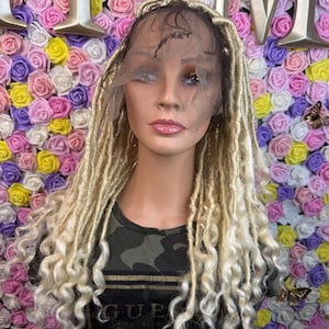 Könnte beinhalten: Ein Schaufensterpuppenkopf mit langen, blonden Dreadlocks. Die Perücke hat eine Spitze vorne und ist mit lockeren Locken an den Enden versehen. Die Schaufensterpuppe trägt ein Camouflage-Print-Shirt mit dem Wort "GUES". Der Hintergrund ist eine Wand aus bunten Rosen.