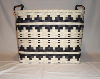 Könnte beinhalten: Ein rechteckiger geflochtener Korb mit einem sich wiederholenden geometrischen Muster in Schwarz und Creme. Der Korb besteht aus flachen, ineinandergreifenden Streifen und verfügt über zwei schwarze geflochtene Griffe am oberen Rand.
