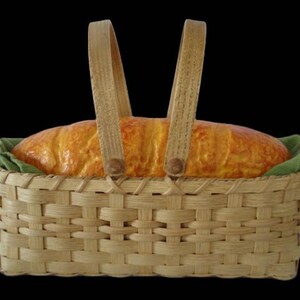 K&ouml;nnte beinhalten: Ein rechteckiger, geflochtener Korb aus hellbraunem Holz mit zwei aufrechten Griffen. Im Korb liegt ein goldbrauner Brotlaib auf einem gefalteten, olivgr&uuml;nen Stoffeinsatz. Der Korb weist ein klassisches Flechtmuster auf.