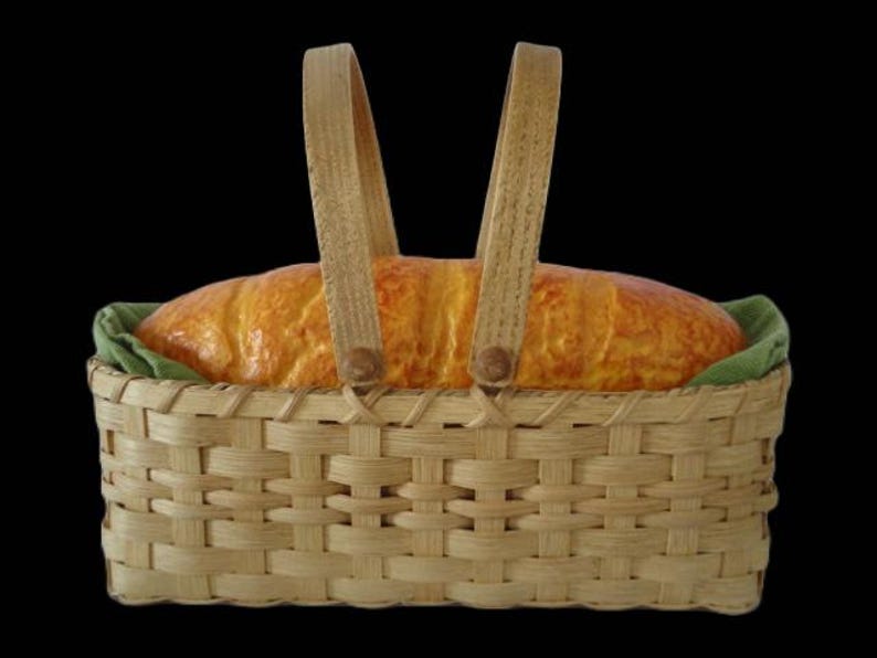 K&ouml;nnte beinhalten: Ein rechteckiger, geflochtener Korb aus hellbraunem Holz mit zwei aufrechten Griffen. Im Korb liegt ein goldbrauner Brotlaib auf einem gefalteten, olivgr&uuml;nen Stoffeinsatz. Der Korb weist ein klassisches Flechtmuster auf.