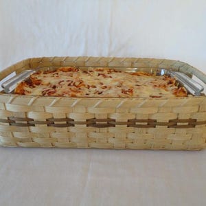 May include: A rectangular woven basket with light tan and dark brown horizontal accents, featuring two integrated handles. Inside the basket sits a clear glass rectangular baking dish filled with a baked pasta dish topped with melted cheese.