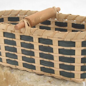 May include: A rectangular woven basket featuring a pattern of natural light tan and black splints. The basket has a sturdy, smooth, cylindrical wooden handle attached to the top rim, extending across the center of the open top section.