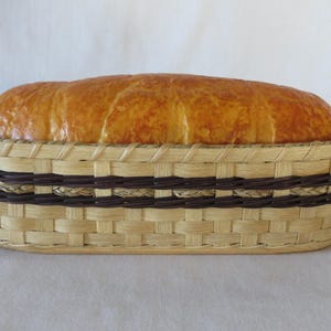 May include: A golden brown loaf of bread sits inside an oval woven basket. The basket features a light tan natural reed base with two horizontal bands of dark brown dyed reed woven around the centre, creating a distinct contrast in colour.