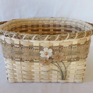 May include: An oval woven basket made of light-colored wood splints with a braided rim and two side handles. A strip of tan burlap wraps around the upper section, decorated with three fabric flowers featuring small wooden beads at their centers.
