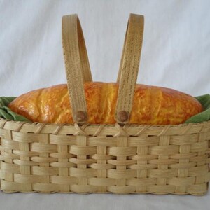 May include: A rectangular woven light-colored wood basket with two upright handles. Inside the basket sits a golden-brown loaf of bread resting on a green fabric liner. The basket features a classic over-under weave pattern and sturdy construction.