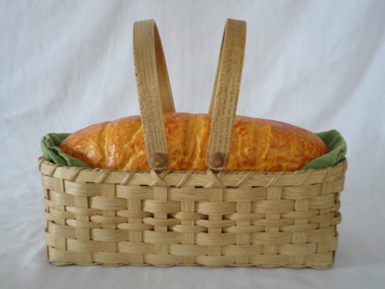 K&ouml;nnte beinhalten: Ein rechteckiger, geflochtener Korb aus hellem Holz mit zwei aufrechten Griffen. Im Korb liegt ein goldbrauner Brotlaib auf einem gr&uuml;nen Stofffutter. Der Korb zeichnet sich durch ein klassisches Flechtmuster und eine stabile Konstruktion aus.