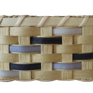Puede incluir: Una cesta rectangular tejida con una base de fibras naturales en tono bronceado claro, con acentos horizontales intercalados en negro sólido y lavanda suave. La estructura consiste en tiras planas entrelazadas con un borde superior reforzado.