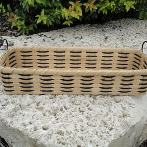 May include: A rectangular woven basket featuring light tan natural reed material with dark brown accent strips woven throughout the sides. The basket includes two black metal handles with a curved, decorative design attached to each end.