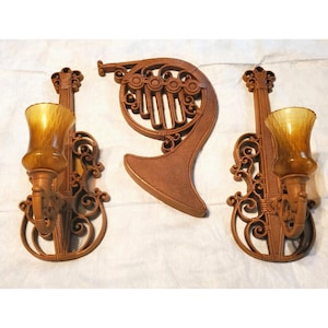 May include: A set of three wall-mounted decorative pieces in a brown finish. Two pieces are violin-shaped sconces with amber-colored glass shades, and the third piece is a central horn-shaped wall plaque with a textured surface and scrollwork.