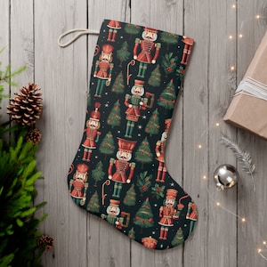 May include: Christmas stocking with a festive pattern of red and green nutcrackers and Christmas trees on a dark background. The stocking has a cream-colored loop for hanging and is made of fabric.