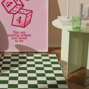 K&ouml;nnte beinhalten: Ein rosa gerahmtes Poster mit einer Grafik von W&uuml;rfeln und dem Text "You are exactly where you need to be". Das Poster h&auml;ngt an einer Wand &uuml;ber einem gr&uuml;n-wei&szlig; karierten Teppich.