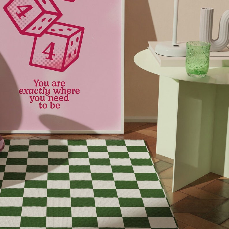 K&ouml;nnte beinhalten: Ein rosa gerahmtes Poster mit einer Grafik von W&uuml;rfeln und dem Text "You are exactly where you need to be". Das Poster h&auml;ngt an einer Wand &uuml;ber einem gr&uuml;n-wei&szlig; karierten Teppich.