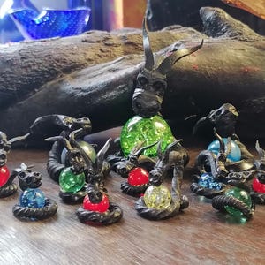Könnte beinhalten: Eine Sammlung kleiner schwarzer Metalldrachenfiguren, die farbige Glasmurmeln halten. Die Drachen sind auf einer Holzoberfläche angeordnet.
