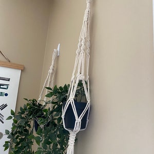 Pode incluir: Dois suportes de macram&eacute; para plantas com uma planta verde em um deles. Os suportes s&atilde;o feitos de corda de algod&atilde;o branco natural e est&atilde;o pendurados em ganchos brancos em uma parede bege.
