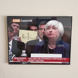 May include: A television screen displaying a news report with the headline "YELLEN: I'M STRONGLY OPPOSED TO AUDIT THE FED". The report features a woman with white hair speaking, and a man holding a yellow sign that reads "Buy Bitcoin".