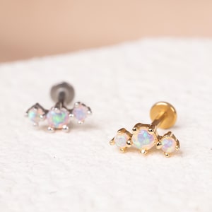 May include: Two flat back stud earrings featuring a trio of prong-set round opals. One earring has a silver-toned metal post and setting, while the other has a gold-toned metal post and setting. Each piece displays three iridescent stones.
