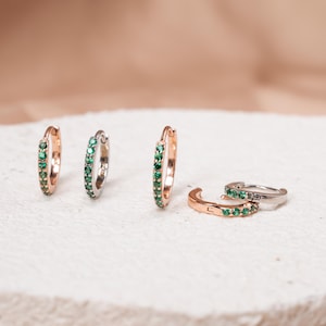 May include: Several small hoop earrings in rose gold and silver, each adorned with a row of small, vibrant green gemstones. The earrings are displayed on a textured, off-white surface.