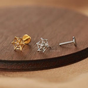 May include: Three silver and gold spider web earrings with a small clear stone in the center. The earrings are on a wooden surface.