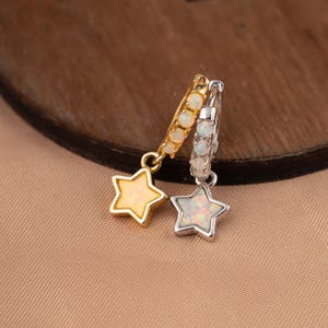 Puede incluir: Dos pendientes de aro con colgantes en forma de estrella. Un aro es de tono dorado con una estrella dorada, y el otro es de tono plateado con una estrella plateada. Ambos aros y estrellas están incrustados con piedras blancas iridiscentes.