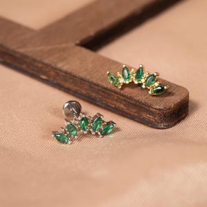 May include: Two curved stud earrings featuring marquise-cut green stones arranged in a fan shape. Each earring is accented with small clear stones set in a metallic frame. One earring is gold-toned, and the other is silver-toned with a flat back post.