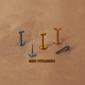 May include: Four titanium body jewellery pieces with square tops in gold, silver, and rose gold. The jewellery is laid out on a brown fabric surface. The text "G23 Titanium" is printed on the surface.