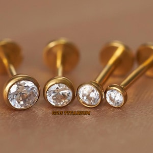 Peut inclure: Cinq bijoux de corps en titane ton or avec des pierres précieuses rondes transparentes. Les bijoux sont pour les piercings et sont étiquetés "G23 Titanium".