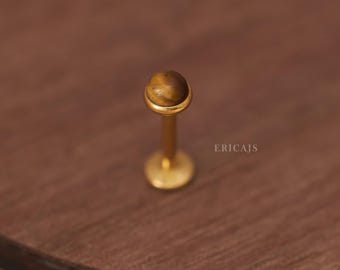 May include: Gold body jewelry with a tiger's eye gemstone. The jewelry is a labret stud with a flat back.