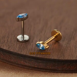 May include: Two titanium body jewellery pieces with blue marquise-shaped gemstones. The piece on the left is silver and the piece on the right is gold. The text "G23 Titanium" is printed on the wooden surface.