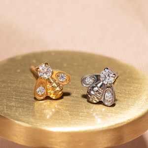 May include: Two bee-shaped stud earrings, one gold-toned and one silver-toned, each adorned with sparkling clear gemstones. The earrings are displayed on a gold-colored surface, showcasing their intricate design and elegant appeal.