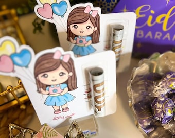 May include: Two cards with cartoon girl illustrations holding heart-shaped balloons. Each card has a small tube and Arabic text. A purple box with gold text and a blue star pattern is in the background. A decorative bowl with purple candies is also visible.