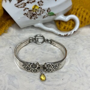 May include: A silver bracelet crafted from repurposed silverware, featuring floral detailing and a toggle clasp. A yellow teardrop-shaped charm dangles from the center. The bracelet is displayed near a white teacup.