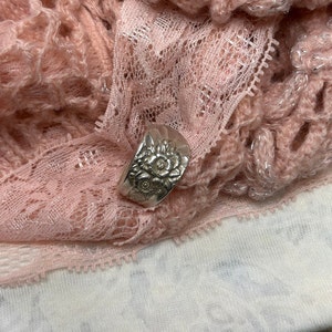 May include: A silver ring with a floral design. The ring is on a pink lace background.
