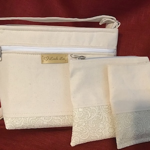 Könnte beinhalten: Drei weiße Stofftaschen mit Spitzenbesatz. Die größte Tasche hat einen Reißverschluss und ein goldenes Etikett mit dem Text "Linda Lee".