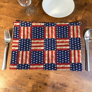 Op de afbeelding: Een rechthoekige placemat met een patchwork-ontwerp van de Amerikaanse vlag. De placemat heeft afwisselende vierkanten van blauw met witte sterren en rood-witte strepen. Een vork, lepel, bord en wijnglas zijn zichtbaar.