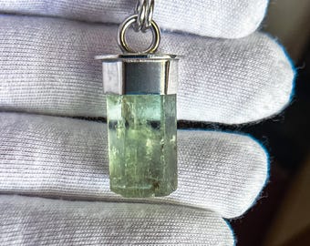 May include: A pendant featuring a raw, hexagonal, light green crystal gemstone set in a polished silver-toned metal cap. The crystal has a translucent appearance with visible internal inclusions and is attached to a thin silver-toned chain.
