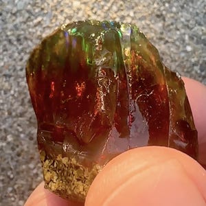 May include: A rough, natural, green and red opal gemstone with iridescent flashes of color. The opal is a raw, unpolished stone with a rough, irregular shape.