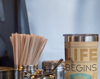 May include: A stainless steel travel mug with a lid, featuring the text "LIFE BEGINS after COFFEE" in a vintage style. The mug has a beige background with a cityscape design and is displayed on a black surface.