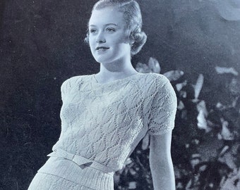 May include: A black and white photo of a woman wearing a knitted dress with a lace pattern. The dress has a fitted bodice and a flowing skirt. The woman is standing in front of a leafy background.
