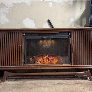 Könnte beinhalten: Elektrischer Kamin aus dunklem Holz. Der Kamin hat eine Glasfront mit simuliertem Feuereffekt. Der Schrank hat vertikale Lamellen an den Seiten und einen geschwungenen Sockel. Die Breite beträgt ca. 150 cm.