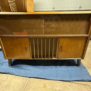 Puede incluir: Gabinete de madera vintage con acabado marrón claro. El gabinete presenta una cubierta de altavoz con diseño de diamantes, dos puertas pequeñas y una rejilla metálica dorada. Se apoya sobre cuatro patas cortas.