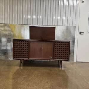 Puede incluir: Una consola estéreo moderna de mediados de siglo en un cálido acabado marrón. La consola presenta una tapa abatible, dos puertas y rejillas de altavoz con un patrón geométrico. Se apoya sobre patas anguladas.