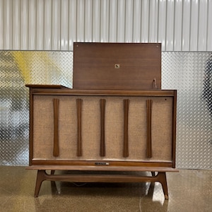 Könnte beinhalten: Eine Stereoanlage aus Holz im Mid-Century-Stil mit einer gewebten Frontplatte und einem Klappdeckel. Die Konsole hat eine dunkle Holzoberfläche und steht auf abgewinkelten Beinen. Die Frontplatte hat vertikale Holzakzente.