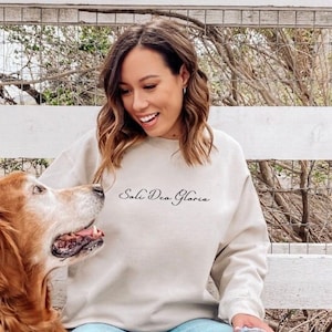 May include: A woman wearing a beige sweatshirt with the text "Soli Deo Gloria" in black script, sits on a wooden deck with a golden retriever dog. The woman is smiling and looking at the dog.