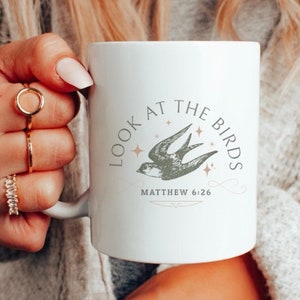 May include: White ceramic mug with the text "LOOK AT THE BIRDS", a bird illustration, and Matthew 6:26. The mug has a white handle. The mug is held by a person wearing rings and a grey sweater.