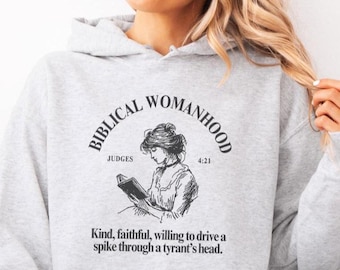 May include: A pale grey hooded sweatshirt with a front pocket and a drawstring hood. The sweatshirt displays a black graphic with the words "BIBLICAL WOMANHOOD" and an illustration of a woman reading. The text "JUDGES 4:21" is also present.