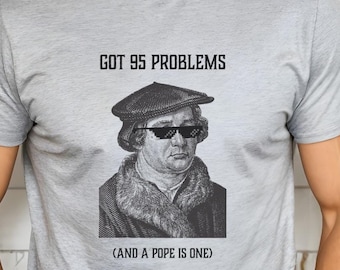 May include: A gray t-shirt with a black and white image of Martin Luther wearing sunglasses and a hat. The text on the shirt reads "GOT 95 PROBLEMS (AND A POPE IS ONE)"